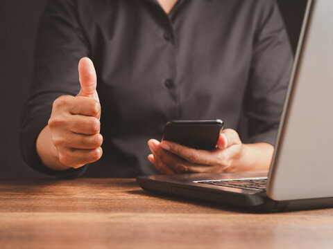 Person Giving Thumbs Up While Holding Smartphone by Laptop