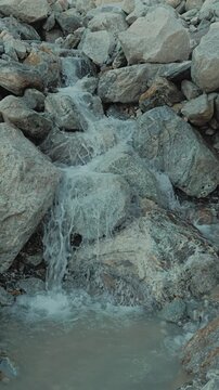 Water flows dynamically over rough gray rocks in a mountain stream. Under soft natural light. Cool tones predominate, and the texture of the wet rocks resembles white foam. Slow motion