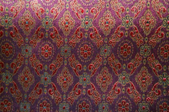 A piece of traditional songket fabric featuring a maroon background and intricate gold thread (zari) weaving, forming a dense, luxurious floral-geometric pattern.