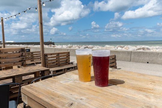 Two artisanal craft beers on wooden deck in outdoor cafe, near ocean at Mers-les-Bains, Normandy, golden pale ale and dark amber ale served at beachfront bar with Atlantic waves in the background