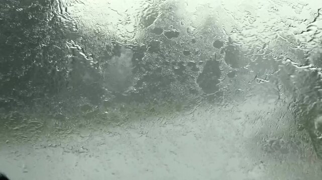Rain covered window with dense droplets and smeared streaks blurring dark trees and faint outdoor shapes, creating muted overcast scene with soft diffused light and calm gloomy mood