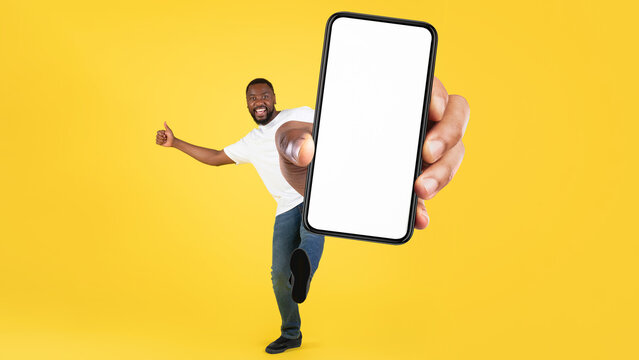 Smiling Black Male Standing Against Yellow Background, Enthusiastically Holding Smartphone With Blank Screen, Presenting New App Or Website In Bright Office Setting. Business Concept.