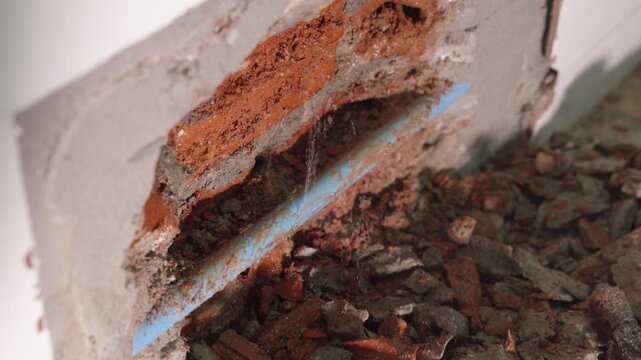Close-up of a leaking water pipe inside a damaged wall, showing plumbing failure, moisture, cracked plaster, water damage, corrosion, and the need for urgent home maintenance and repair.