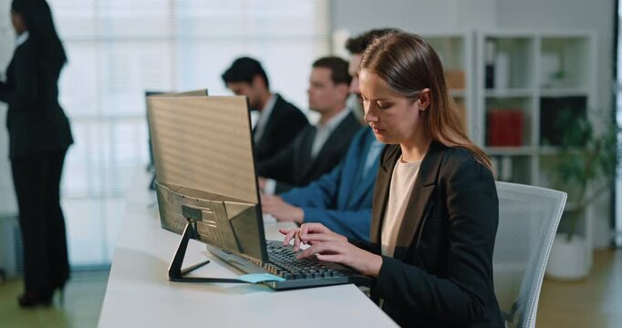 Portrait of Caucasian businesswoman types on keyboard at desktop computer in open office. Focused woman works on company task as colleagues continue routine