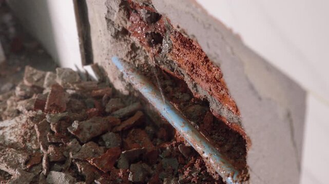 Close-up of a leaking water pipe inside a damaged wall, showing plumbing failure, moisture, cracked plaster, water damage, corrosion, and the need for urgent home maintenance and repair.