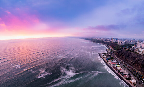 Aerial view of the Lima coastline at sunset, featuring dramatic cliffs, the Pacific Ocean, and the city skyline along the highway in Lima, Callao Region, Peru.