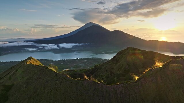 Aerial view of Mount Batur and Lake Batur at sunrise with volcanic craters, lush green slopes, and golden light in Bali, Indonesia.