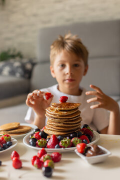 Sweet toddler child, boy, eating american pancakes with strawberries, cherries and blueberries, topped with syrup and drinking fresh orange juice