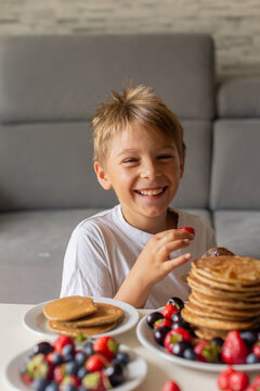 Sweet toddler child, boy, eating american pancakes with strawberries, cherries and blueberries, topped with syrup and drinking fresh orange juice
