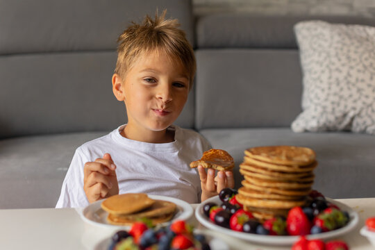 Sweet toddler child, boy, eating american pancakes with strawberries, cherries and blueberries, topped with syrup and drinking fresh orange juice