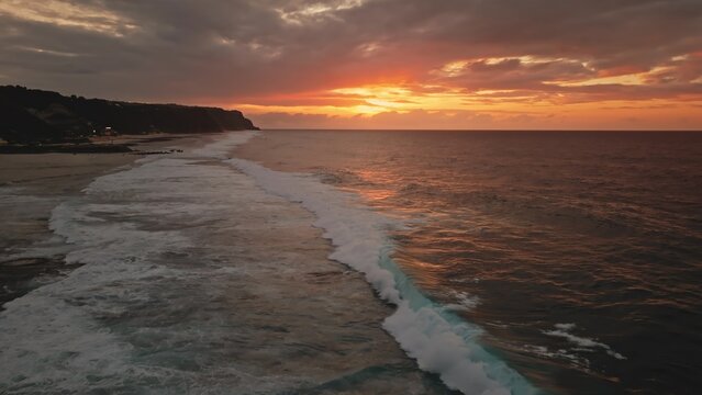 Aerial view of waves crashing against the coastline at sunset with dark cliffs and golden sky in Bali, Indonesia.