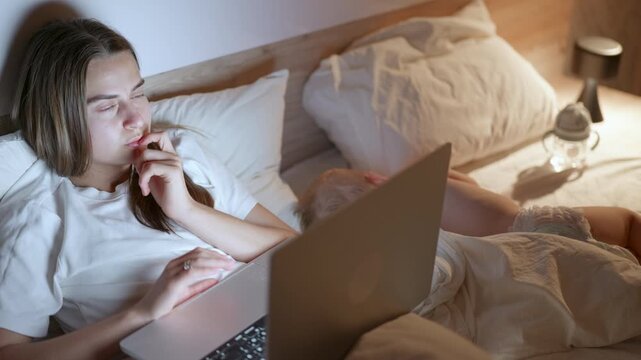 Young woman with long brown hair in white shirt focused on laptop while sitting in bed beside sleeping child in cozy bedroom with soft lighting and bedding