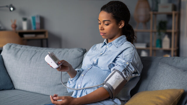 Focused Black Woman In Pregnancy, Measuring Blood Pressure Using A Device At Home, Ensuring Health During Pregnancy, Sitting Comfortably On Sofa In Living Room, Healthcare Concept.