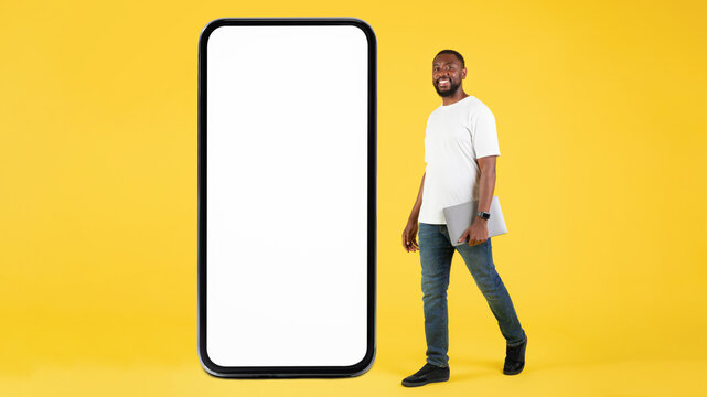 Smiling Black Man Holding Laptop And Walking Beside Large Smartphone With Empty Screen In Studio Setting, Showcasing Technology Concept For Advertisement Or Branding. Mockup Presentation.