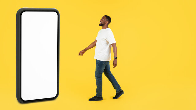 Happy Black Man Smiling While Walking Towards Empty Smartphone Screen In Studio Setting. Bright Yellow Background Enhances The Focus On Modern Technology Concept And Digital Innovation.