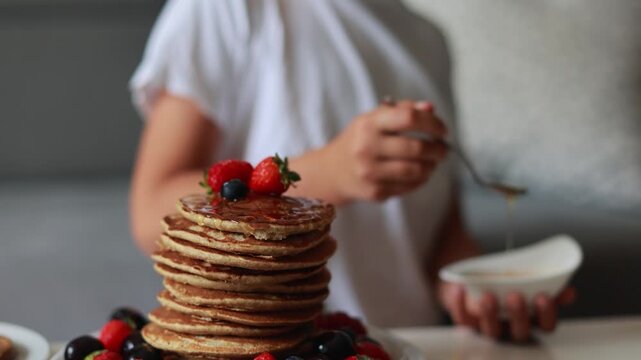 Sweet toddler child, boy, eating american pancakes with strawberries, cherries and blueberries, topped with syrup and drinking fresh orange juice