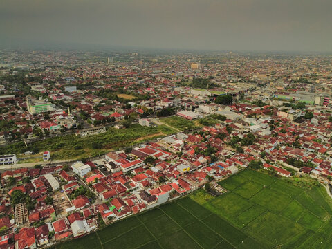 Aerial view of the city of Purwokerto featuring dense residential areas with red rooftops, lush green rice fields, and urban infrastructure in Purwokerto, Central Java, Indonesia.