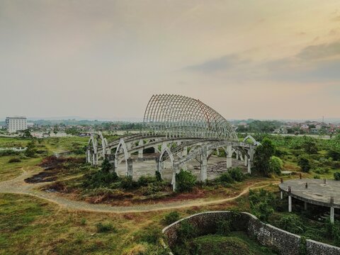 Aerial view of the unfinished Masjid Agung Purwokerto with its distinctive crescent-shaped roof frame, set amidst open grassy fields in Purwokerto, Central Java, Indonesia.