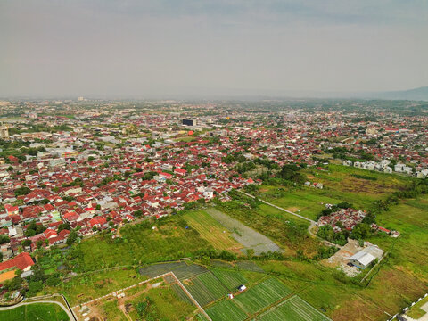 Aerial view of the city of Purwokerto with dense residential neighborhoods featuring red-tiled roofs, surrounded by lush green agricultural fields in Purwokerto, Central Java, Indonesia.