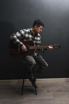 Hombre adulto con camisa a cuadros tocando la guitarra ac&uacute;stica sentado en un taburete sobre fondo oscuro en estudio.
