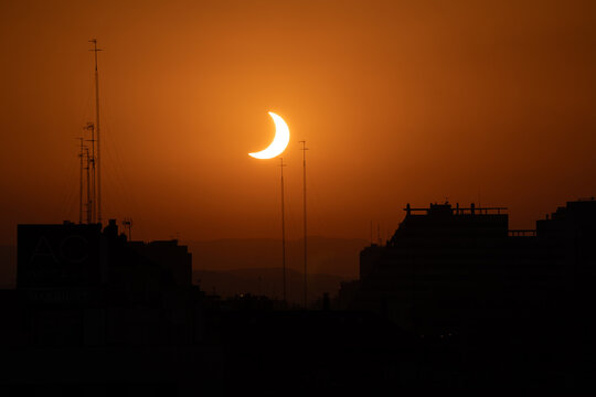Crescent sun of a partial solar eclipse sinks toward city rooftops, dark building silhouette, tall antenna mast, guy wires, deep orange sky, hazy mountain ridge behind the city