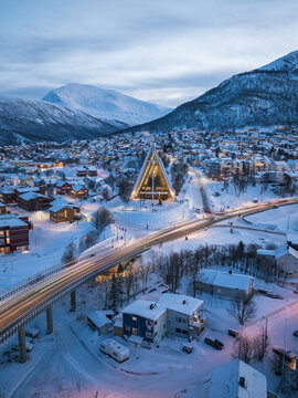 Aerial view of the Arctic Cathedral illuminated at dusk, surrounded by snow-covered houses, a bridge with light trails, and mountains in Tromso, Troms, Norway.
