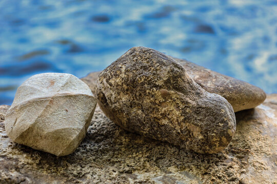 Several large rounded stones lie on a rocky surface near a body of water. One of them features an unusual geometric shape that stands out against the brownish texture of other boulders.