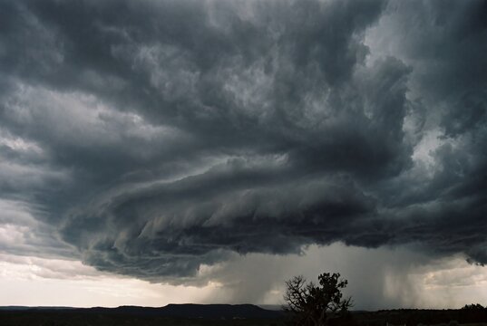 Dark storm clouds gather over a landscape, creating a dramatic sky. Rain is visible in the distance, with a silhouette of trees in the foreground.