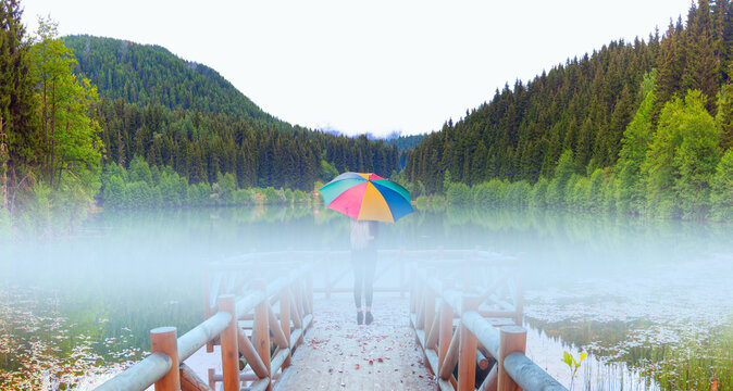 A beautiful blonde woman with a multicolored umbrella stands on the pier amidst the fog, watching her surroundings - Morning evaporation of water over the lake - Amazing autumn landscape with Karagol 