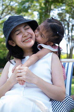Little girl kissing her smiling mother on the cheek in a sunny park, expressing family love, affection, happiness, parenting, childhood, emotional bonding, outdoor lifestyle, precious family moments.