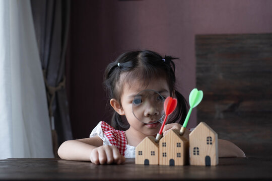Little girl using a magnifying glass to inspect wooden house models with target darts, symbolizing property research, investment decisions, home buying, financial literacy, goal setting.