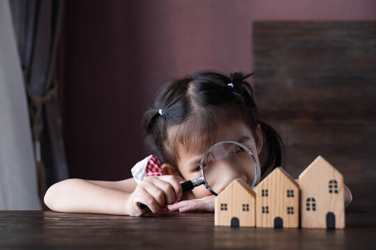 Little girl examining wooden house models with a magnifying glass, symbolizing property research, home inspection, real estate investment, childhood learning, curiosity, and future financial planning.