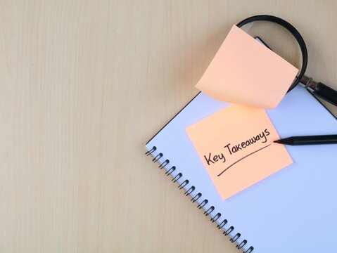 Key Takeaways text handwritten on adhesive note placed on book with pen and magnifying glass on wooden background. Business summary, learning concept, and analysis top view with copy space