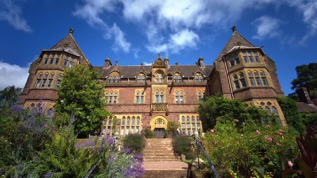 Victorian High Gothic revival facade of Knightshayes Court framed by vibrant purple flowers in the estate gardens, photographed in Tiverton, Devon, England, on 24.08.2024.