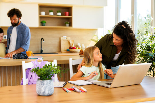 Mother assisting daughter with online schoolwork on laptop while father helps son in the background promoting education and family bonding