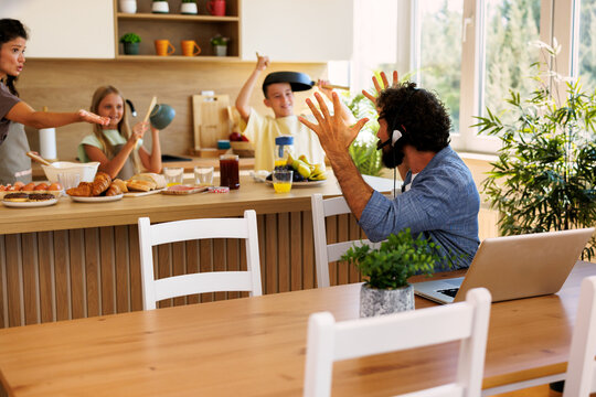 Frustrated father working on a laptop in a kitchen with loud children making noise with kitchen utensils