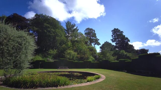 A tranquil wide shot of a circular lily pond surrounded by manicured lawns and high yew hedges at Knightshayes Court in Tiverton, Devon, United Kingdom, filmed on 24.08.2024