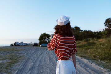 Obraz premium Back view of a woman in striped shirt and skirt along a dirt road by the beach, capturing lifestyle, outdoor travel and casual summer fashion.