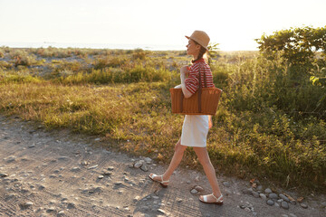 Obraz premium Walking woman in a straw hat crosses a sunlit path, wearing a striped top and white skirt while carrying a woven basket outdoors in a golden hour field