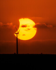 Fototapeta premium Partial solar eclipse behind a wind turbine silhouette during sunset, combining renewable energy concept with astronomical phenomenon