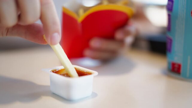 Close-up of hand dipping crispy golden French fry into red ketchup sauce, with box of fried potatoes blurred in background. Fast food, snack and casual dining concept.