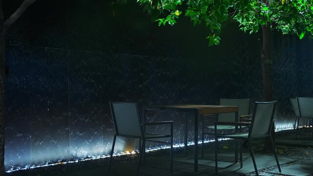 Outdoor restaurant terrace with empty chairs and tables by illuminated flowing water wall, Nighttime cafe seating under tree in decorative garden setting with artificial waterfall 4k