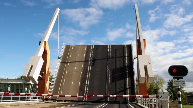 Dutch drawbridge lowers over roadway beneath blue sky, with barriers and red signal controlling traffic. Sign text Keep marked road sections clear warning drivers near movable bridge crossing