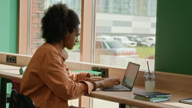 African American young adult typing on laptop while sitting by window in modern library. Clip is ideal for promoting online education, remote work, or student lifestyle campaigns