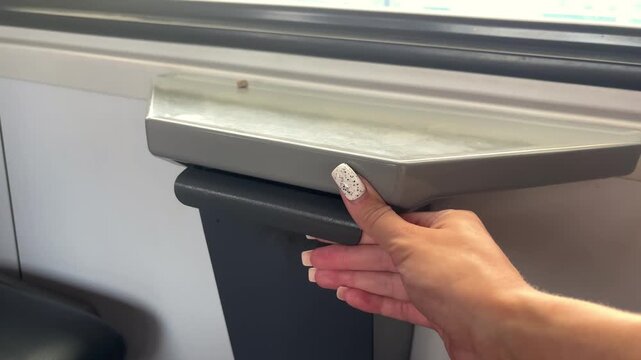 Woman opening a small waste compartment below a train window, commuter rail interior, cleanliness, travel convenience, public transport.