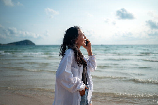 Happy young woman talking on her smartphone at a tropical beach, enjoying vacation, remote work
