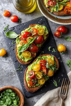 Overhead view of two avocado toasts topped with cherry tomatoes basil and olive oil on dark slate board