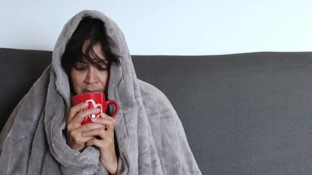 Sick middle eastern mature woman wrapped in a blanket, shivering and drinking a hot beverage .influenza virus symptoms.