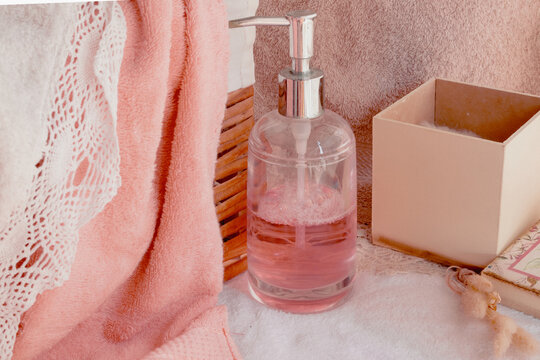 Bathroom countertop featuring a pump bottle with pink liquid soap. Soft peach towel. White embroidered towel. And decorative boxes
