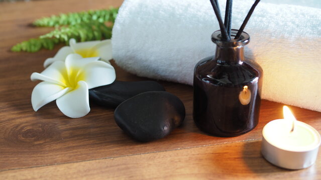 Spa arrangement with white plumeria flowers, black massage stones, reed diffuser bottle, burning candle light, and rolled towel on wooden surface.